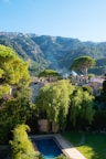 A serene rural landscape featuring a villa surrounded by lush greenery and tall trees in the foreground, with a swimming pool visible in the yard. In the background, there are majestic mountains partially covered with dense forests, and the sky is clear and blue.