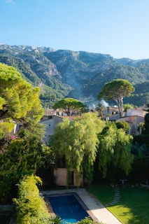 A serene rural landscape featuring a villa surrounded by lush greenery and tall trees in the foreground, with a swimming pool visible in the yard. In the background, there are majestic mountains partially covered with dense forests, and the sky is clear and blue.