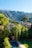 A serene rural landscape featuring a villa surrounded by lush greenery and tall trees in the foreground, with a swimming pool visible in the yard. In the background, there are majestic mountains partially covered with dense forests, and the sky is clear and blue.