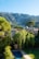 A serene rural landscape featuring a villa surrounded by lush greenery and tall trees in the foreground, with a swimming pool visible in the yard. In the background, there are majestic mountains partially covered with dense forests, and the sky is clear and blue.