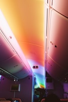 A colorful airplane cabin interior featuring overhead storage compartments and a ceiling illuminated with a gradient of hues ranging from pink, orange, and yellow to blue and green. The cabin's lighting creates a soothing and modern atmosphere, highlighting details such as seatback screens and an illuminated exit sign.