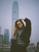 A fashionable person poses against an urban backdrop featuring tall skyscrapers and a Ferris wheel in the distance. They wear glasses, a grey sweater, and a dark, textured coat. The atmosphere suggests a cloudy day.