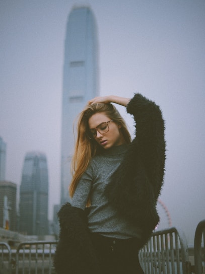 A fashionable person poses against an urban backdrop featuring tall skyscrapers and a Ferris wheel in the distance. They wear glasses, a grey sweater, and a dark, textured coat. The atmosphere suggests a cloudy day.