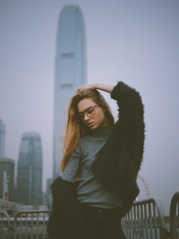 A fashionable person poses against an urban backdrop featuring tall skyscrapers and a Ferris wheel in the distance. They wear glasses, a grey sweater, and a dark, textured coat. The atmosphere suggests a cloudy day.