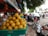 A street market scene with a large green basket filled with yellow lemons prominently displayed in the foreground. The background features a bustling street with people, motorcycles, and cars moving by. A leafy tree canopy provides some shade over the scene, and a blue and red signage with text is partially visible.