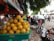A street market scene with a large green basket filled with yellow lemons prominently displayed in the foreground. The background features a bustling street with people, motorcycles, and cars moving by. A leafy tree canopy provides some shade over the scene, and a blue and red signage with text is partially visible.