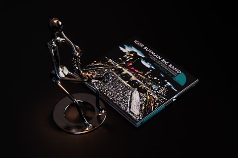 A sleek metallic sculpture of a person playing a saxophone is positioned beside a CD case featuring album art for 'Igor Butman Big Band.' The setting is dark, highlighting the reflective surface of the sculpture and the vibrant colors of the CD cover art.