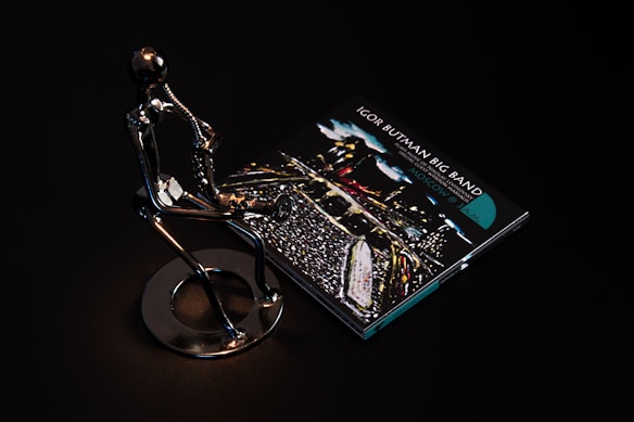 A sleek metallic sculpture of a person playing a saxophone is positioned beside a CD case featuring album art for 'Igor Butman Big Band.' The setting is dark, highlighting the reflective surface of the sculpture and the vibrant colors of the CD cover art.