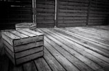 A wooden deck with two wooden crates sitting on top, surrounded by a wooden fence. The texture of the weathered wood is evident, with a monochromatic color palette.