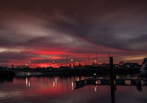 The image captures a serene marina scene during sunset, with dramatic red and orange hues in the sky. Silhouettes of sailboats and yachts are visible against the vibrant sky. Calm water reflects the colors, enhancing the tranquil atmosphere.