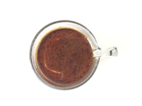 A clear mug filled with dark brown coffee, viewed from above against a white background. The surface of the coffee shows a creamy, slightly frothy texture with small bubbles and speckles, indicating a freshly brewed beverage.