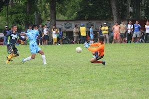 A group of soccer players are actively engaged on a grassy field, with one player in an orange jersey making a sliding save attempt to stop the ball. Spectators stand in the background watching the game.