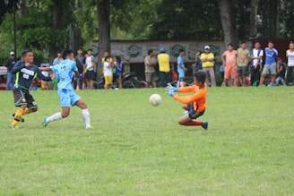 A group of soccer players are actively engaged on a grassy field, with one player in an orange jersey making a sliding save attempt to stop the ball. Spectators stand in the background watching the game.