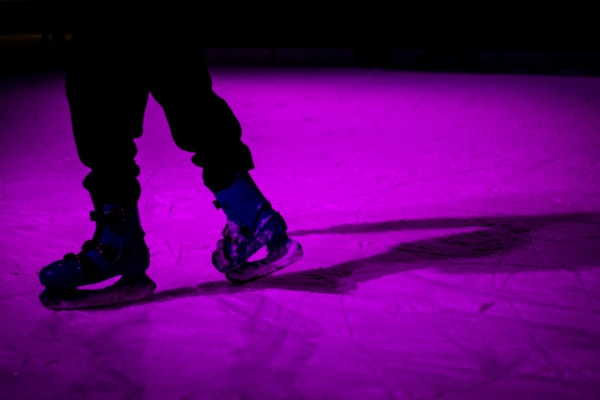 A pair of ice skates on a rink bathed in vibrant purple lighting. The skater's lower legs and skates are visible, casting a dramatic shadow across the ice surface.