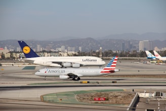 Two commercial airplanes, one from Lufthansa and another from American Airlines, taxi on a runway in an airport. The background features a city skyline with tall buildings and distant hills.