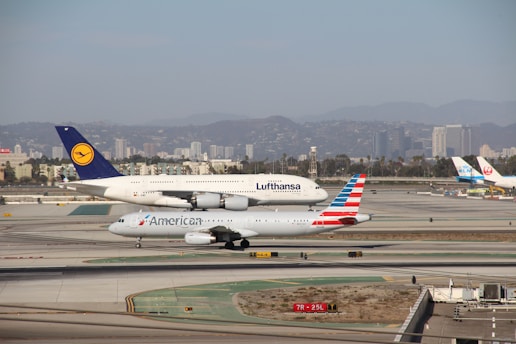 Two commercial airplanes, one from Lufthansa and another from American Airlines, taxi on a runway in an airport. The background features a city skyline with tall buildings and distant hills.