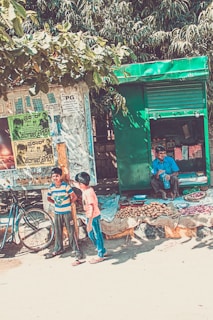 A street scene featuring a small roadside stall with a green shutter, selling potatoes and onions. A man is seated in the stall, drinking from a bottle. In the foreground, two boys are standing next to a bicycle, near a wall covered with posters and advertisements. Trees with green leaves form the backdrop, providing shade.