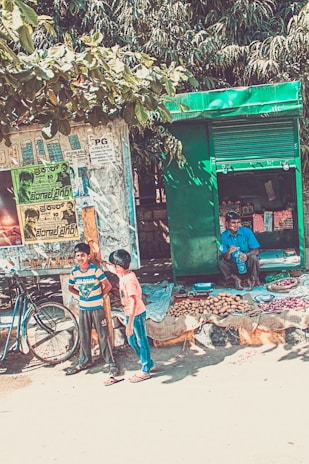 A street scene featuring a small roadside stall with a green shutter, selling potatoes and onions. A man is seated in the stall, drinking from a bottle. In the foreground, two boys are standing next to a bicycle, near a wall covered with posters and advertisements. Trees with green leaves form the backdrop, providing shade.