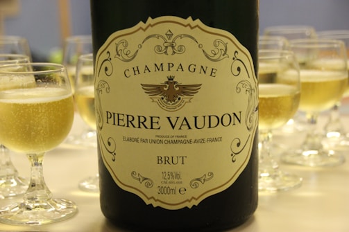 A large bottle of Pierre Vaudon champagne is prominently featured with an ornate label, surrounded by filled wine glasses with a light golden bubbly liquid. The background is softly blurred, emphasizing the focus on the bottle and the glasses.