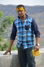 A man with a yellow and red tilak on his forehead is wearing a blue checkered shirt and black glasses. He is standing beside a concrete wall with greenery and hills in the background. He holds a yellow paper in his hand.