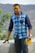 A man with a yellow and red tilak on his forehead is wearing a blue checkered shirt and black glasses. He is standing beside a concrete wall with greenery and hills in the background. He holds a yellow paper in his hand.