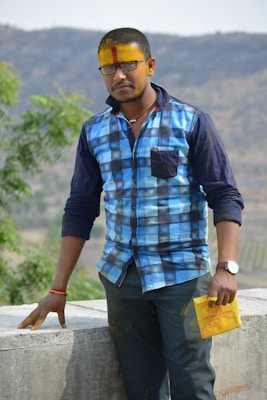A man with a yellow and red tilak on his forehead is wearing a blue checkered shirt and black glasses. He is standing beside a concrete wall with greenery and hills in the background. He holds a yellow paper in his hand.