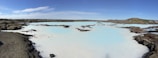 A serene landscape featuring a large geothermal spa with milky blue water surrounded by rugged, dark volcanic rocks under a clear blue sky. Patches of moss can be seen on the rocks, and gentle hills are visible in the distance.