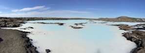 A serene landscape featuring a large geothermal spa with milky blue water surrounded by rugged, dark volcanic rocks under a clear blue sky. Patches of moss can be seen on the rocks, and gentle hills are visible in the distance.