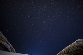 A night sky filled with stars stretches across the image. The top portion of two thatched roofs or huts is visible in the lower corners, suggesting a serene outdoor setting.