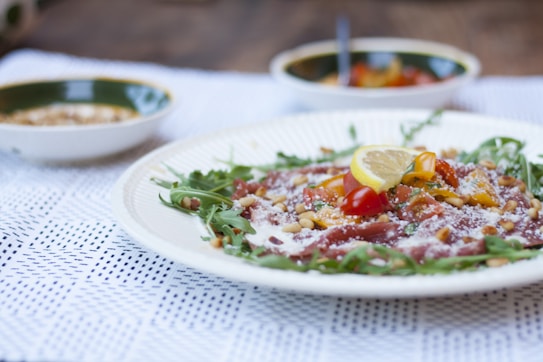 A plate of gourmet salad featuring arugula, cherry tomatoes, lemon slice, pine nuts, and what appears to be a sprinkling of cheese. The salad is presented on a white plate placed on a white, lace-patterned tablecloth. In the background, there are two small bowls, one of which contains a mixture of salad ingredients and a spoon.