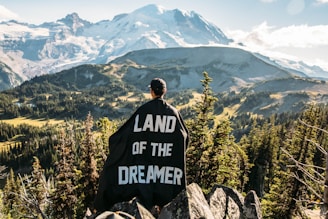 person standing on mountain peak with black land of the dreamer-printed cape on his back