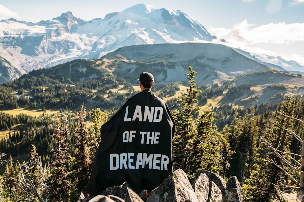 Person, die auf einem Berggipfel mit schwarzem Umhang mit Land of the Dreamer-Aufdruck auf dem Rücken steht