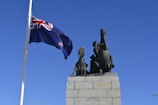 A large stone monument featuring bronze sculptures of two rearing horses and a soldier. A New Zealand flag flutters prominently on a tall flagpole against a clear blue sky.