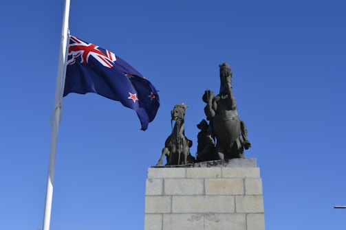 A large stone monument featuring bronze sculptures of two rearing horses and a soldier. A New Zealand flag flutters prominently on a tall flagpole against a clear blue sky.