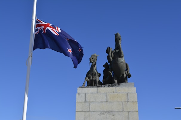 A large stone monument featuring bronze sculptures of two rearing horses and a soldier. A New Zealand flag flutters prominently on a tall flagpole against a clear blue sky.