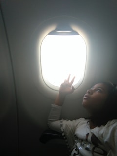 A young girl looks thoughtfully out an airplane window, raising her hand with two fingers extended. Natural light illuminates her face, creating a calm and introspective atmosphere.