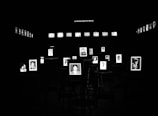 Black and white photographs are displayed prominently in a dark room, each framed and illuminated. They are arranged on stools with additional photos lining the walls in the background.