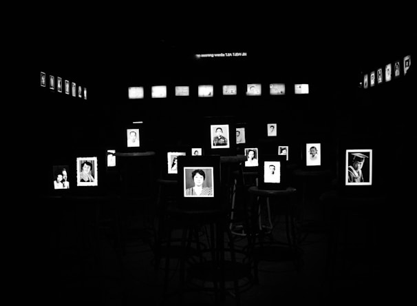 Black and white photographs are displayed prominently in a dark room, each framed and illuminated. They are arranged on stools with additional photos lining the walls in the background.