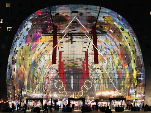 A large, curved building features an expansive glass facade displaying a vibrant, colorful mural. The artwork consists of a mix of abstract shapes and natural elements such as fruits and flowers. Inside, the area is illuminated with a warm glow from various light sources. Red vertical banners hang from the ceiling, and there are decorative circular wreaths with lights near the entrance, where people move about.