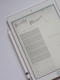 A tablet displaying a digital weekly planner is placed on a light-colored surface. The planner layout includes sections for schedule and events from Monday to Sunday, a to-do list, and a space for notes and gratitude. A stylus rests beside the tablet, suggesting interaction with the planner.
