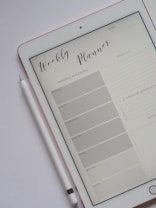 A tablet displaying a digital weekly planner is placed on a light-colored surface. The planner layout includes sections for schedule and events from Monday to Sunday, a to-do list, and a space for notes and gratitude. A stylus rests beside the tablet, suggesting interaction with the planner.