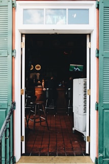 A view through an open doorway leading into a dimly lit bar with a tiled floor. The bar has stools and a small TV showing a sports game. Green shutters frame the door, and an ATM sign is visible above the entrance.