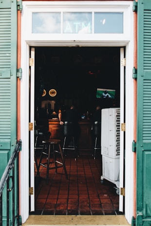 A view through an open doorway leading into a dimly lit bar with a tiled floor. The bar has stools and a small TV showing a sports game. Green shutters frame the door, and an ATM sign is visible above the entrance.