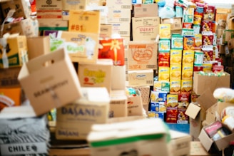 A large assortment of cardboard boxes and packaged goods are stacked and scattered, filling the space with a variety of colors and printed labels. The boxes contain different brands of groceries, with recognizable packaging for items such as snacks and drinks. The arrangement suggests a storage area or supply room filled with non-perishable food items.