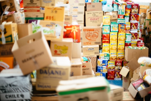 A large assortment of cardboard boxes and packaged goods are stacked and scattered, filling the space with a variety of colors and printed labels. The boxes contain different brands of groceries, with recognizable packaging for items such as snacks and drinks. The arrangement suggests a storage area or supply room filled with non-perishable food items.