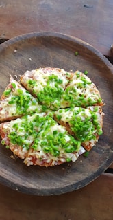 A freshly baked pizza topped with melted cheese and a generous amount of chopped green bell peppers, served on a rustic wooden platter. The pizza is cut into six slices, and sits against a wooden table background.