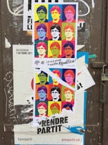 A torn poster featuring a pop art-style collage of a person's face in various colorful squares, similar to Andy Warhol's style. It is placed on a weathered door with other stickers and graffiti. The poster contains multiple colors such as yellow, blue, and red with bold black text at the bottom.