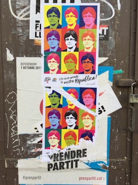 A torn poster featuring a pop art-style collage of a person's face in various colorful squares, similar to Andy Warhol's style. It is placed on a weathered door with other stickers and graffiti. The poster contains multiple colors such as yellow, blue, and red with bold black text at the bottom.