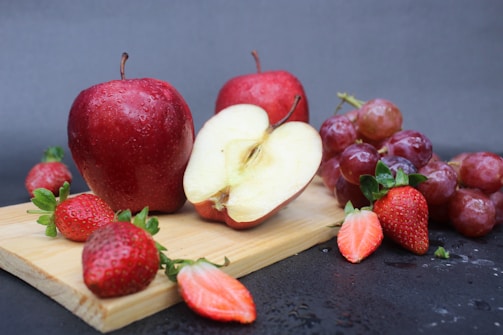 Fresh red apples, halved apples, strawberries, and a bunch of red grapes are arranged on a wooden cutting board. The fruits appear fresh and are laid out against a dark textured background, with some droplets of water visible on the surface.