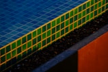 Colorful tiles in green and blue form the edge of a swimming pool, with sunlight creating distinct shadows and highlights. The texture of small pebbles is visible beside the pool.